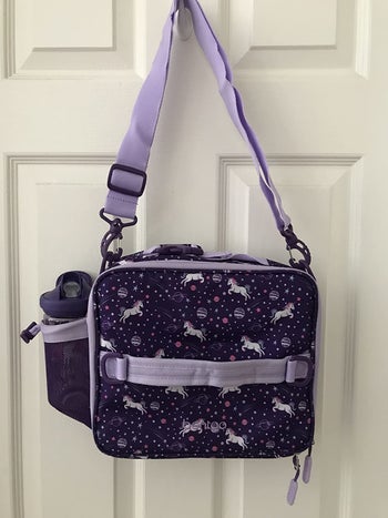 reviewer's photo of the purple bag in unicorn print with a water bottle in the side pocket hanging on a door