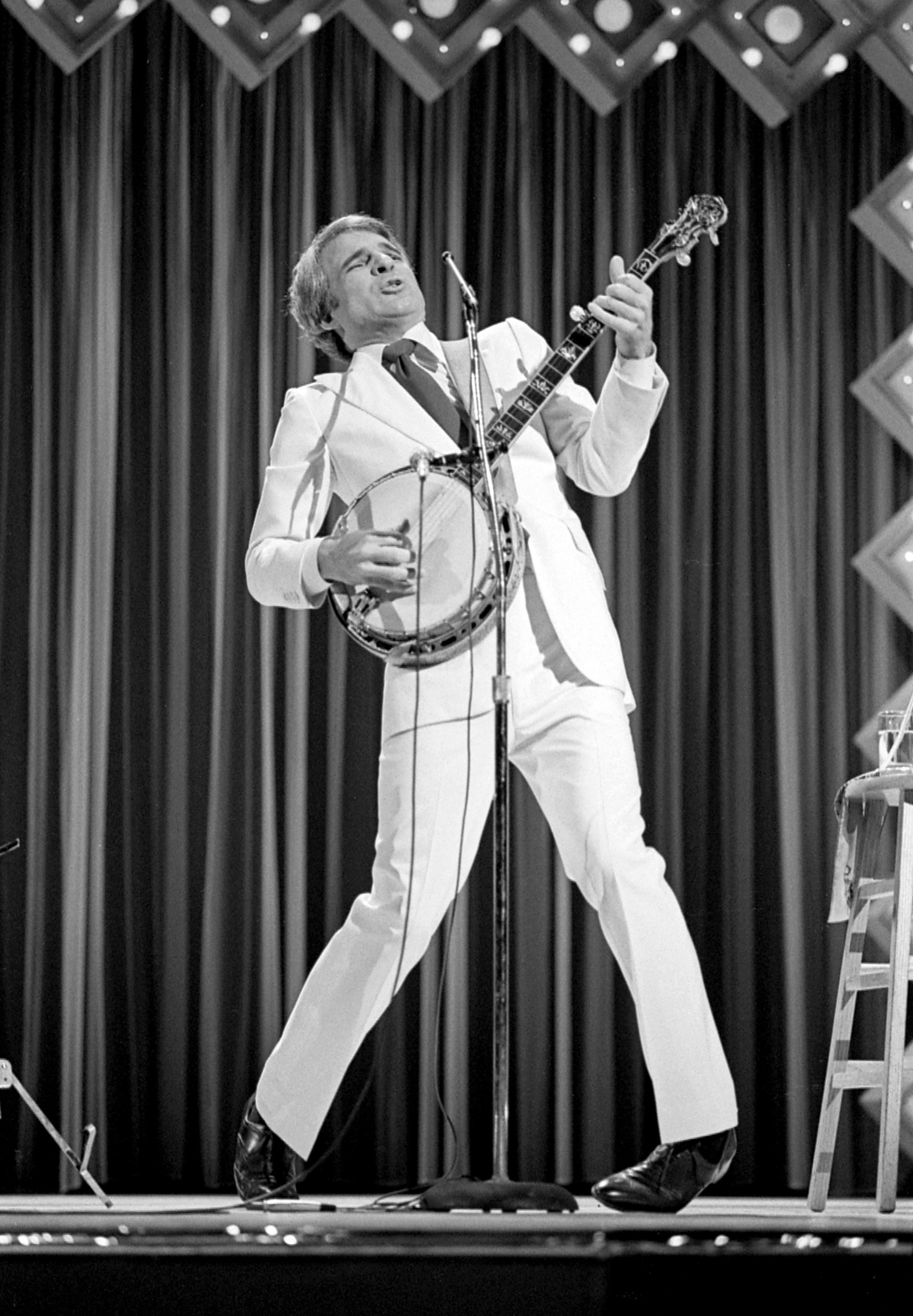 A young Steve Martin performs stand-up onstage while playing a banjo and dancing