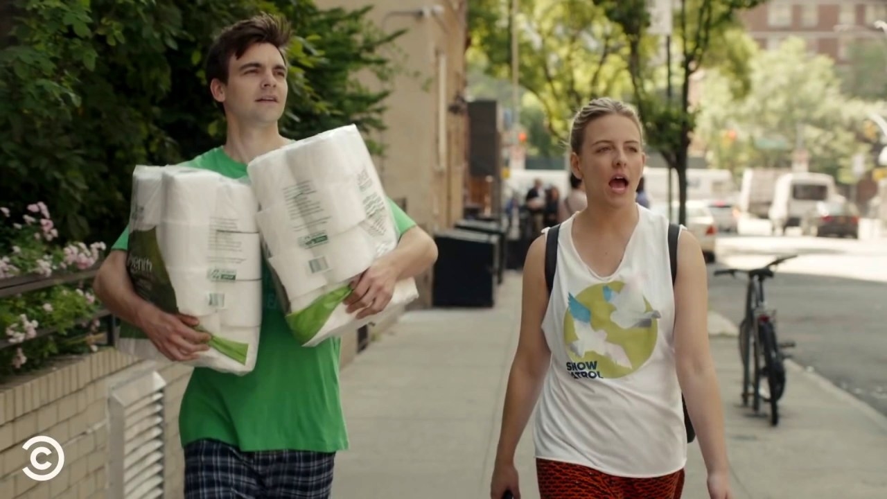 A screenshot of Cary and Brooke walking down the street in the middle of the day, looking a bit of a mess. Cary is in pyjamas, his hair a mess and carrying two large packs of toilet paper, Brooke has her hair tied back and is in gym attire.