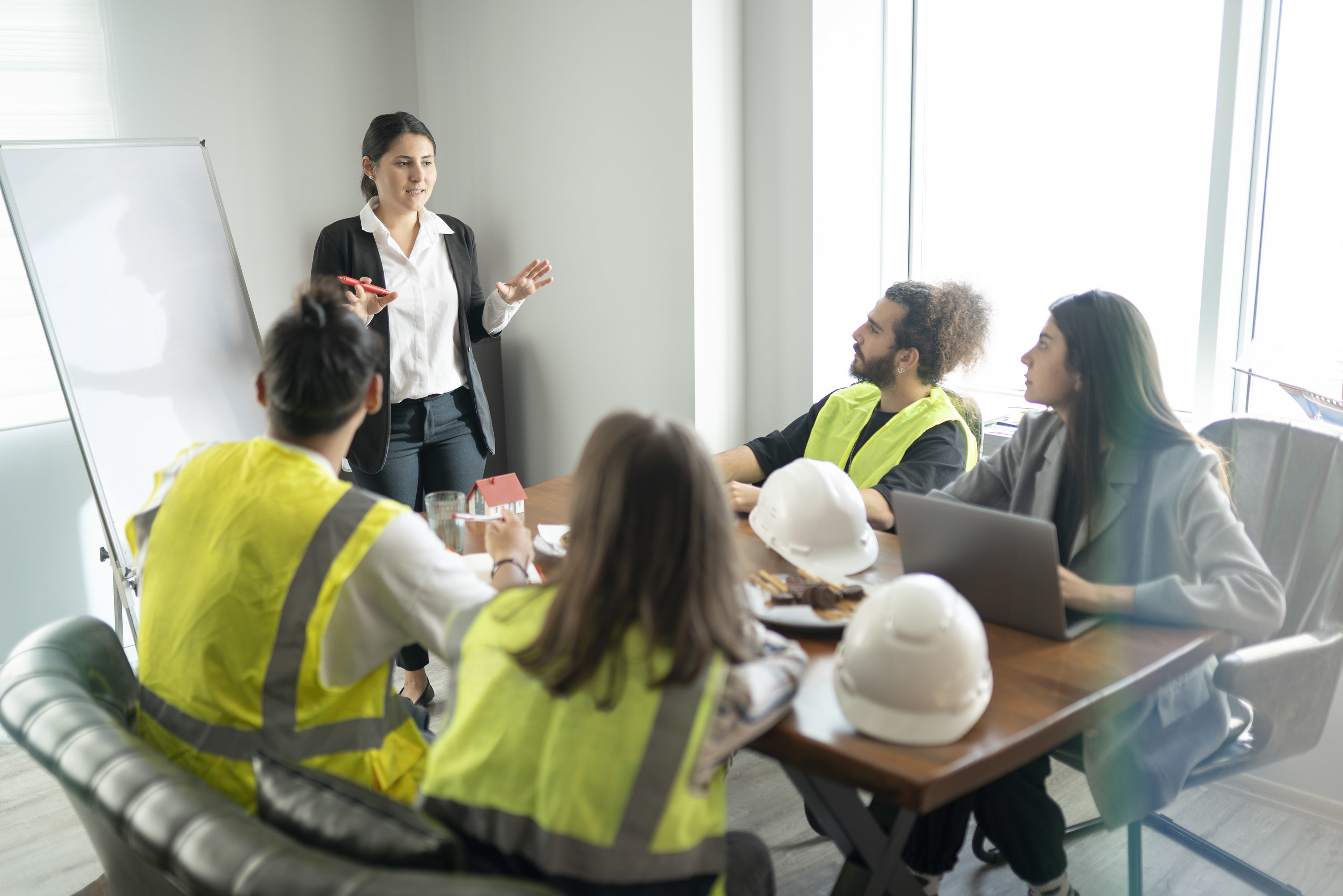 Woman in a blazer giving a presentation to a room of construction workers, some wearing yellow reflective jackets, with hard hats on the table