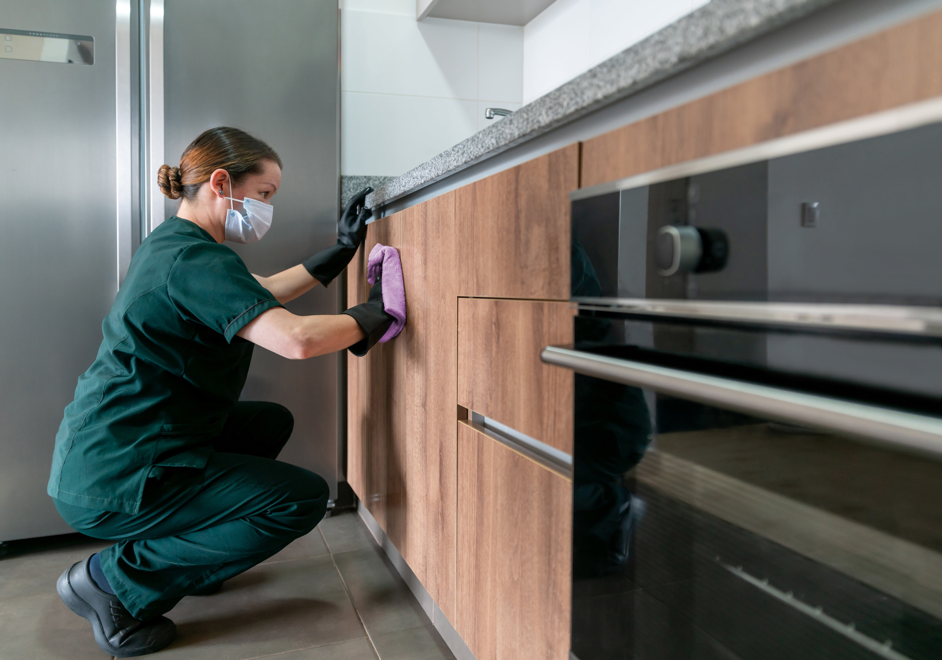Woman wearing a surgical mask and gloves while cleaning the sides of a cabinet in a nice-looking kitchen