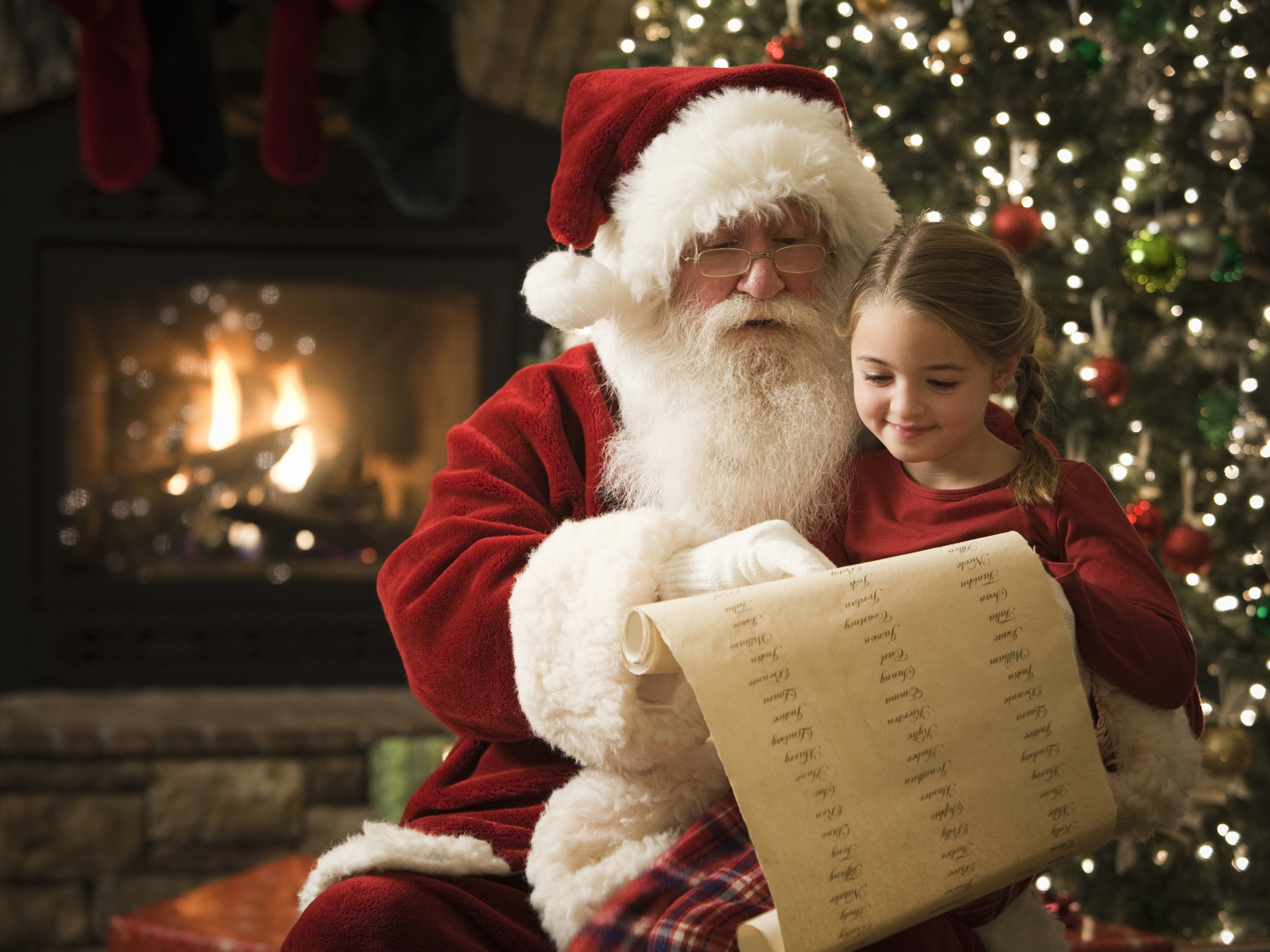 A child sits on Santa&#x27;s lap in front of a Christmas tree and fireplace as Santa points to his list