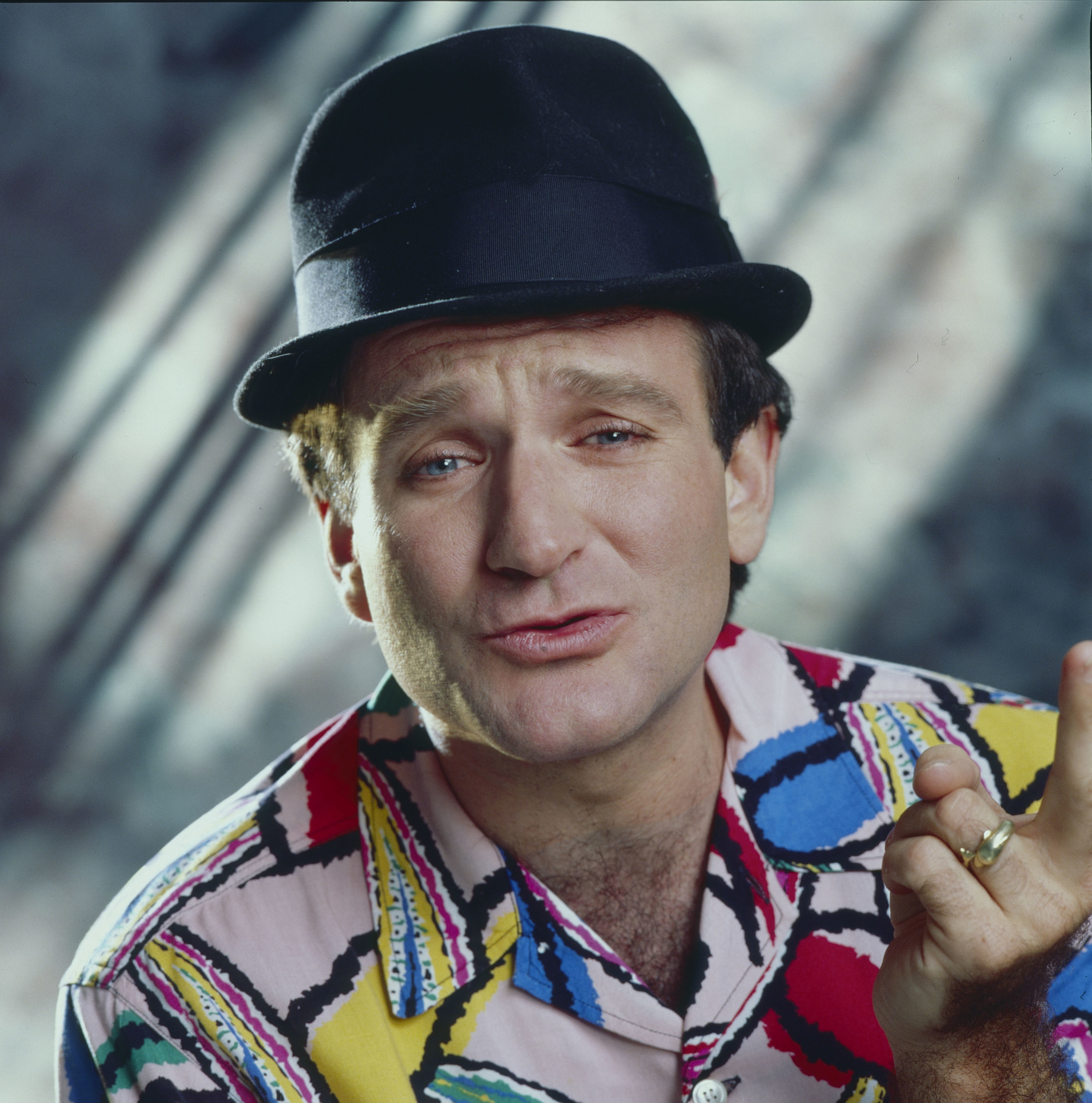 Robin Williams posts wearing a hate while holding up one hand and wearing a pattern shirt