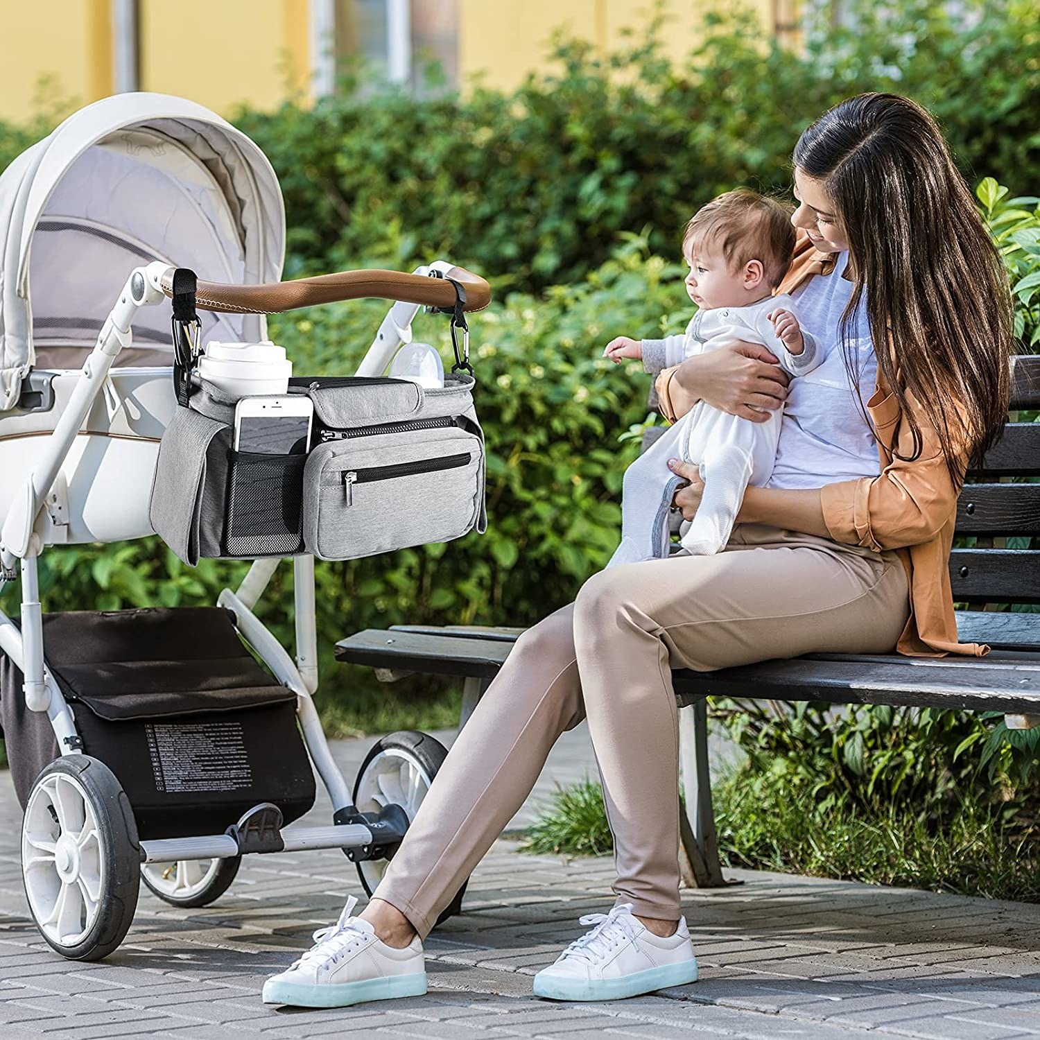a person sitting on a park bench with their baby; the stroller organizer is visible on their stroller