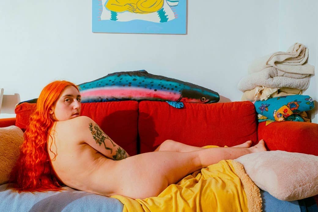 A naked person with an arm tattoo lies on a couch on their side, with their back to the camera, and looks at the camera