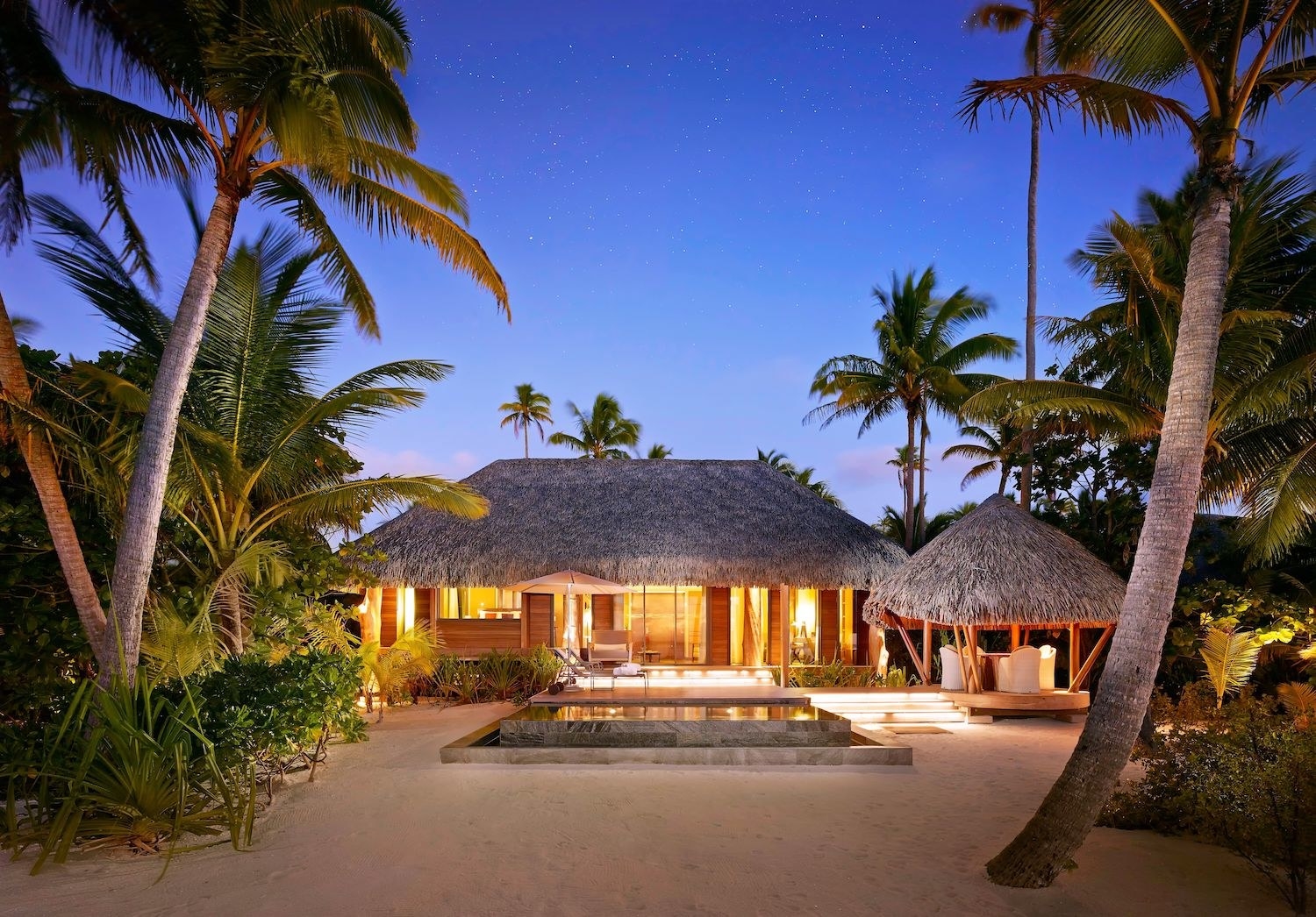 A view of a villa with a thatched roof, a deck with a small plunge pool, and a sandy beach surrounded by palm trees