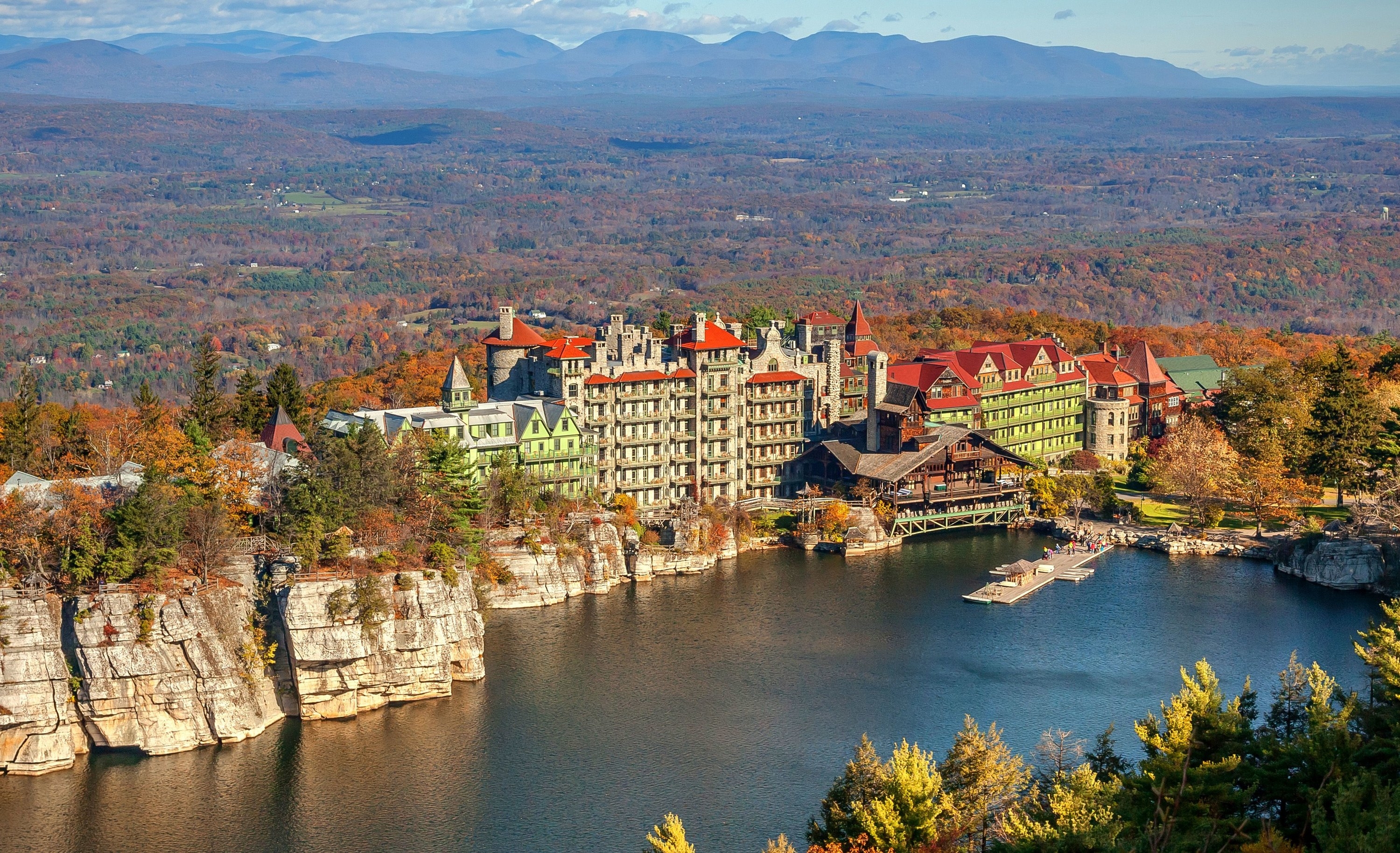 An aerial view of the hotel, which has tall towers set on a cliffside overlooking a lake with rolling hills covered in forest in the background