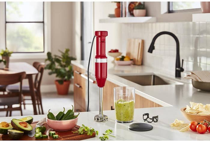 The Kitchenaid immersion blender on a counter next to a cup of pureed food, as well as several jalapenos and halved avocados