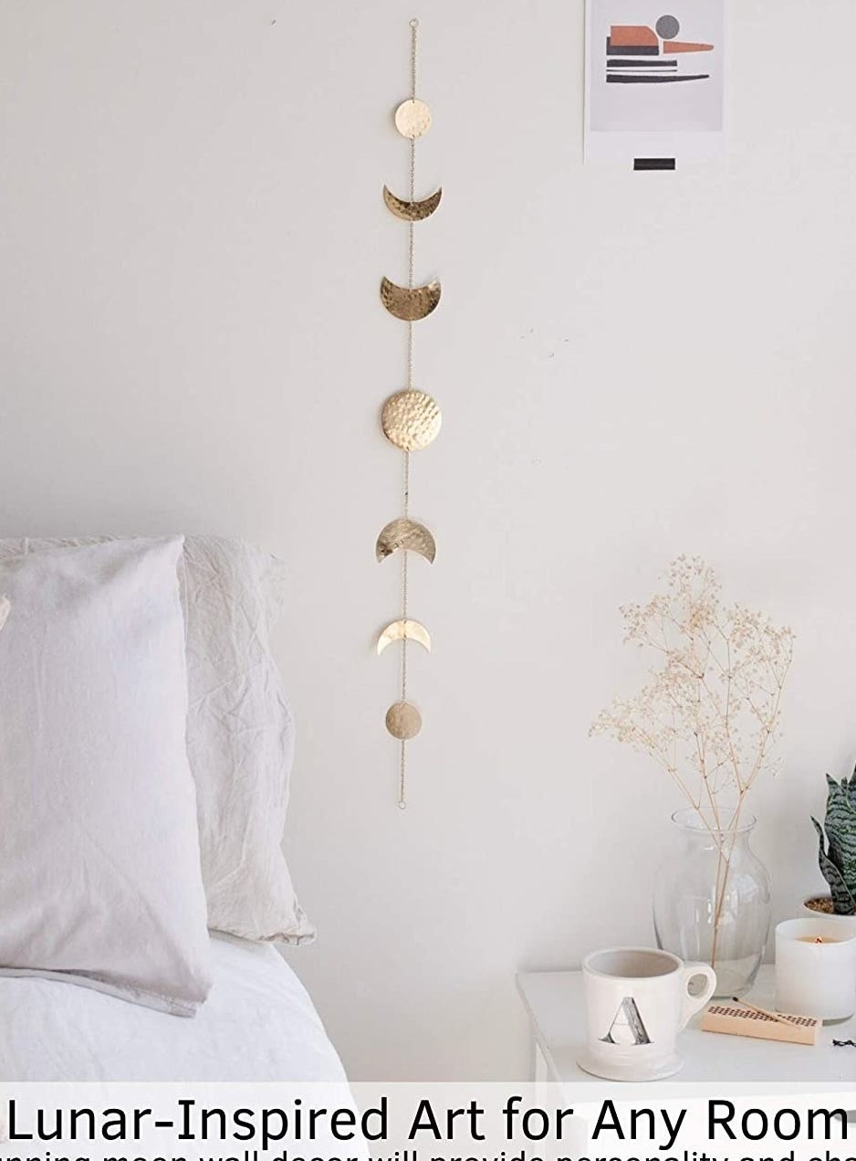 The moon decor hanging on a plain wall above a bed and an end table with a mug and plants on it