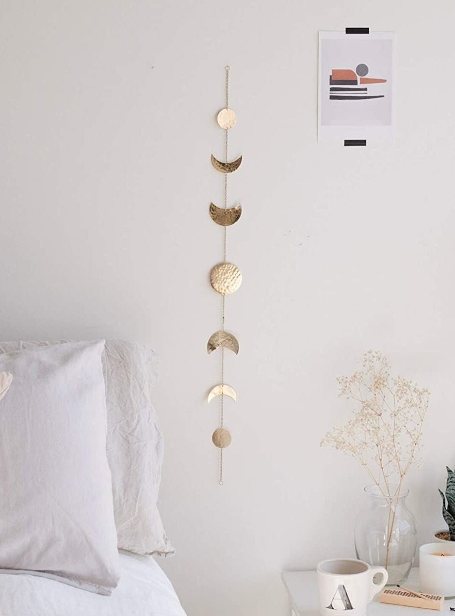 The moon decor hanging on a plain wall above a bed and an end table with a mug and plants on it