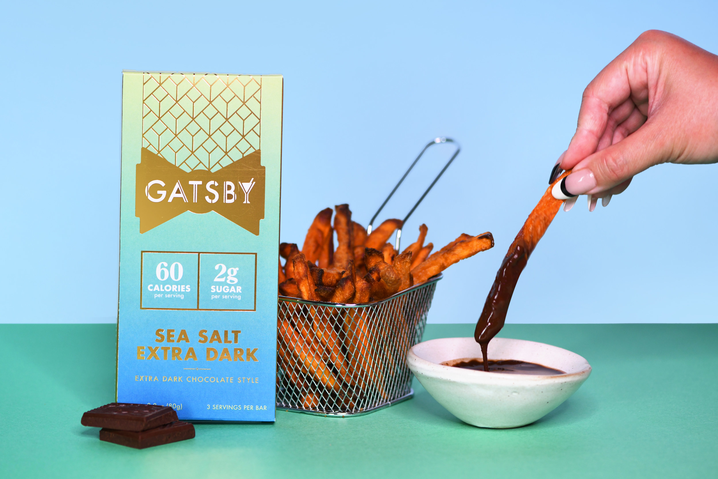 A Gatsby&#x27;s Sea Salt Extra Dark chocolate bar set beside a basket of sweet potato fries and a hand dipping one in a bowl of chocolate