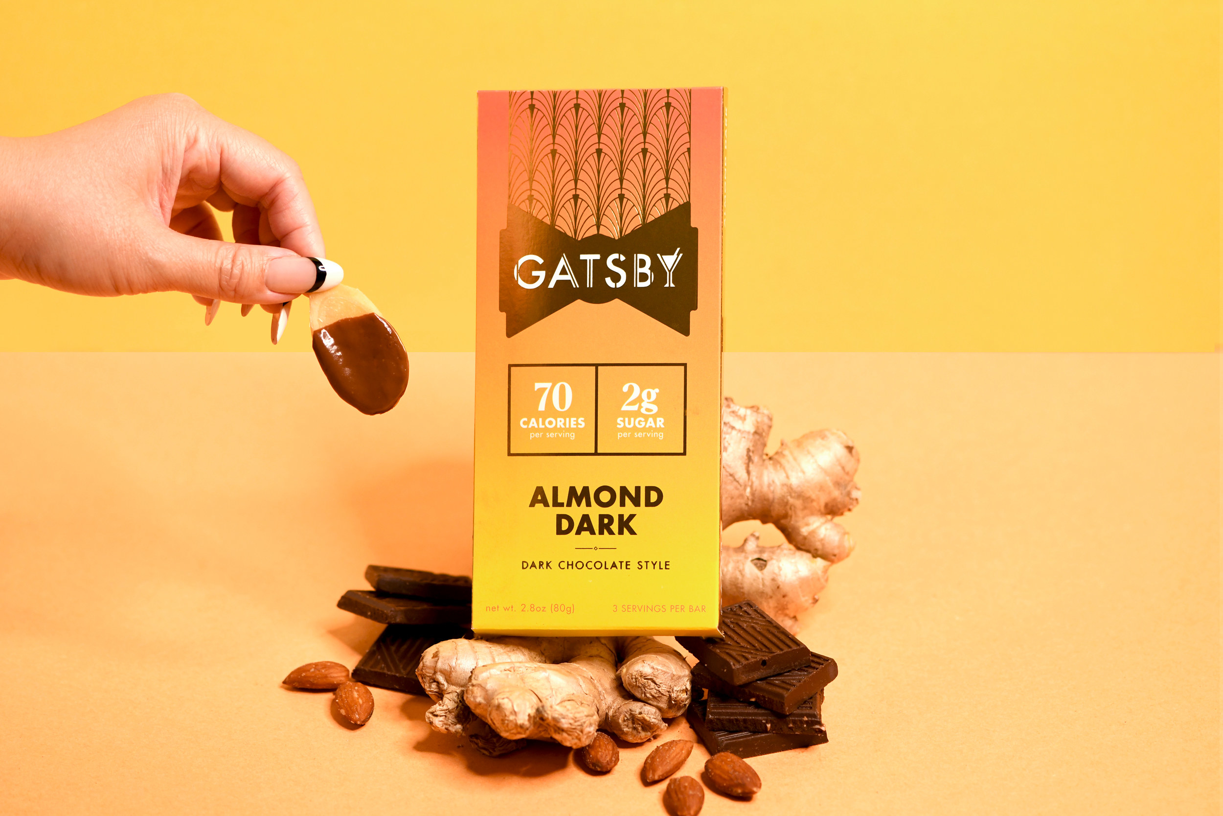 A bar of Gatsby&#x27;s Almond Dark chocolate set on a pile of ginger and chocolate beside a hand holding chocolate dipped ginger