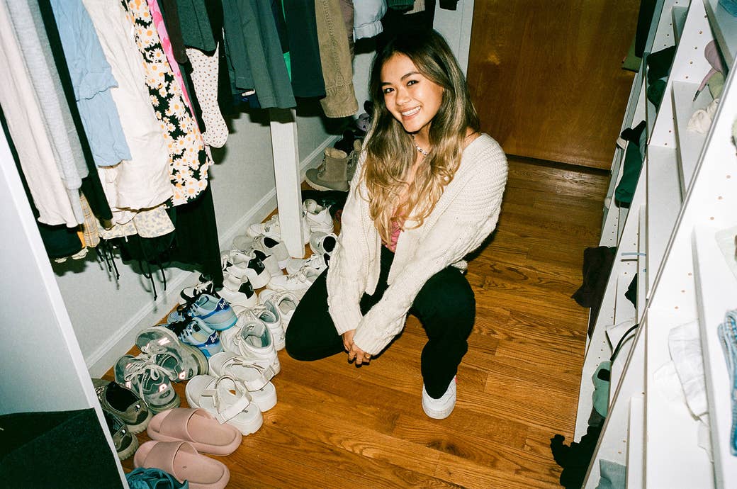 She crouches, smiling, on a wood floor in a closet space next to many pairs of shoes