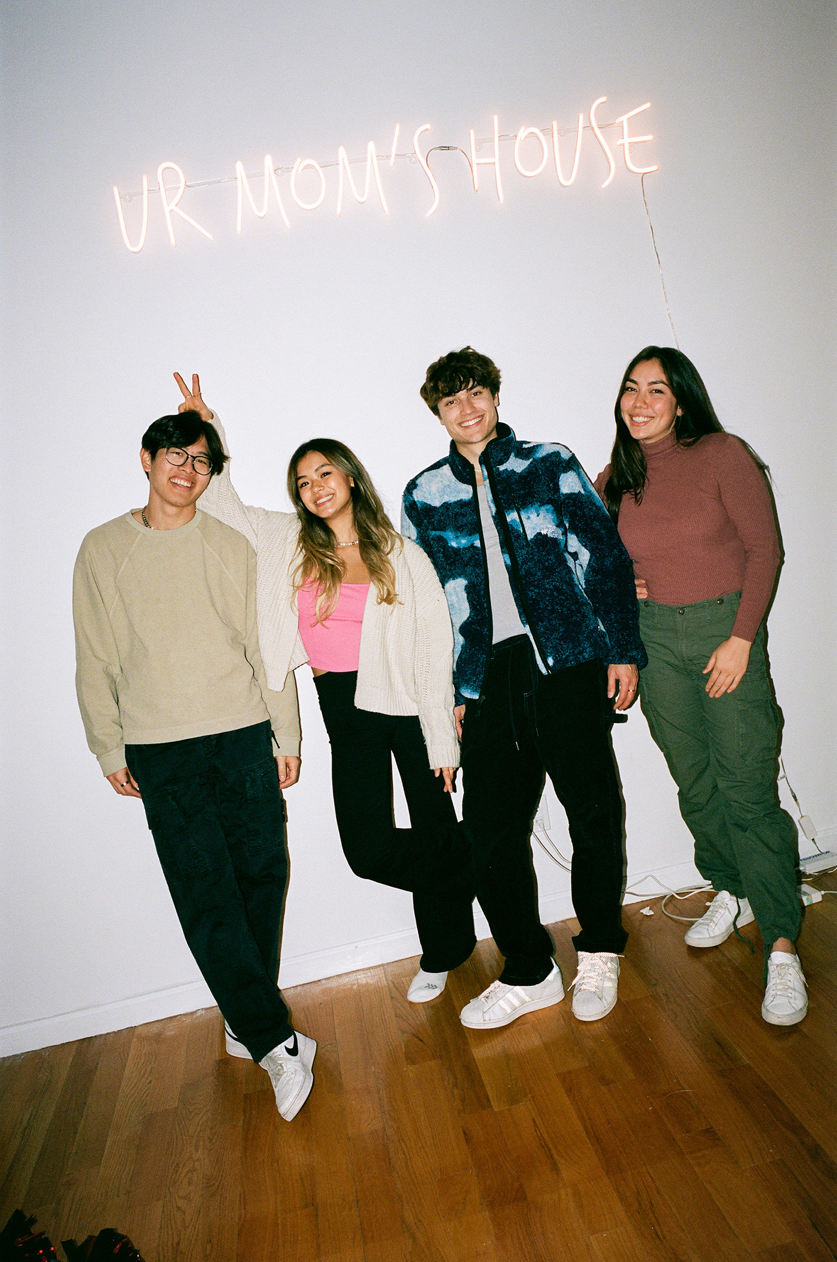 Four smiling people dressed casually stand together on a wood floor, with &quot;Ur Mom&#x27;s House&quot; in lights above them