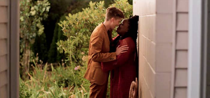 A screenshot from the third season of the show depicts main character Annie's best friend and roommate, Fran, a Black Lesbian in an embrace with her nonbinary partner, Em. Em is smiling, about to kiss Fran up against a white brick wall outdoors.