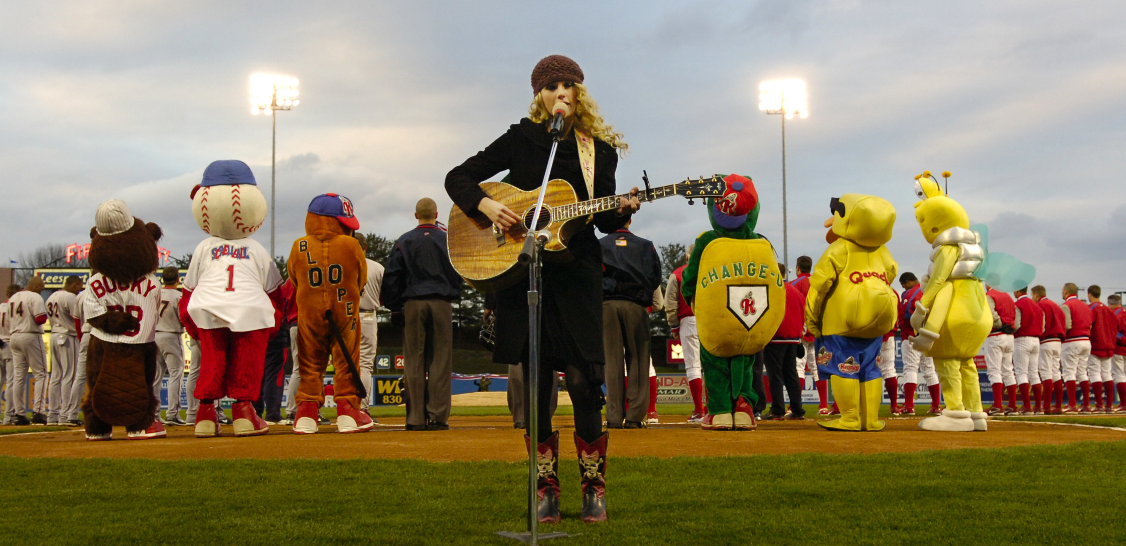 Taylor standing in front of a podium and playing guitar on a grassy field with mascots and team members behind her