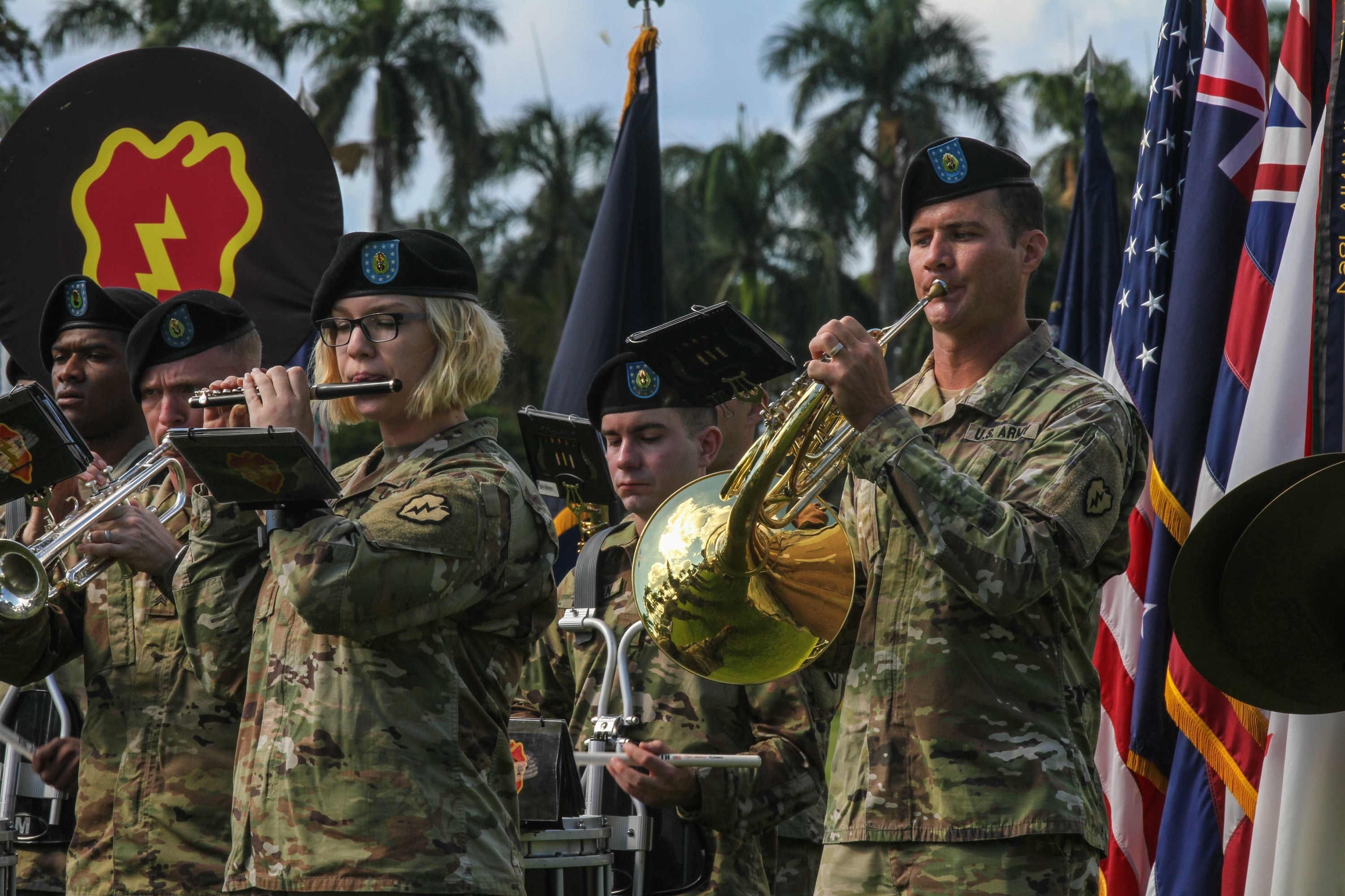 People in Army uniforms playing in a band in front of palm trees and flags
