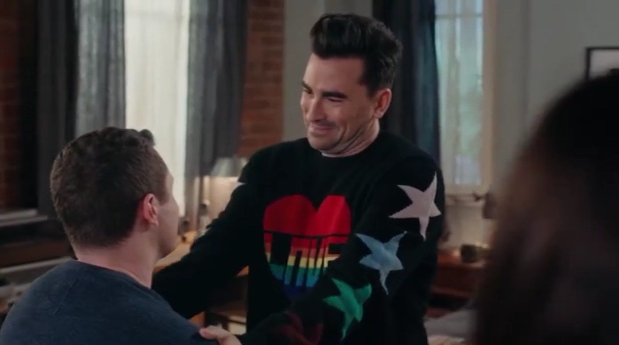 In a screenshot from the season finale, David Rose, wearing a sweater with a rainbow heart with the word &quot;LOVE&quot;, smiles down at his fiancé, Patrick. David has both hands on Patrick&#x27;s shoulders, Patrick is looking up at David, only his profile visible.
