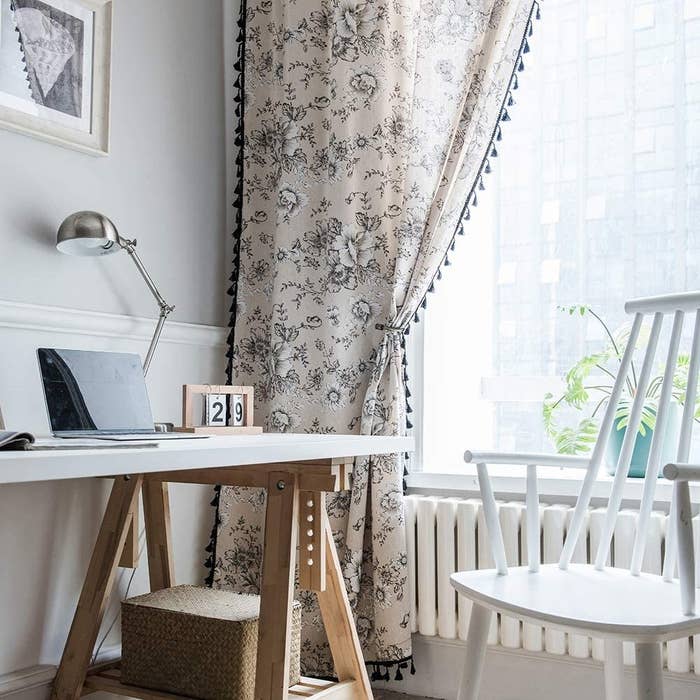 room with a desk beside of a window with a curtain panel that&#x27;s floral print with tassel trim