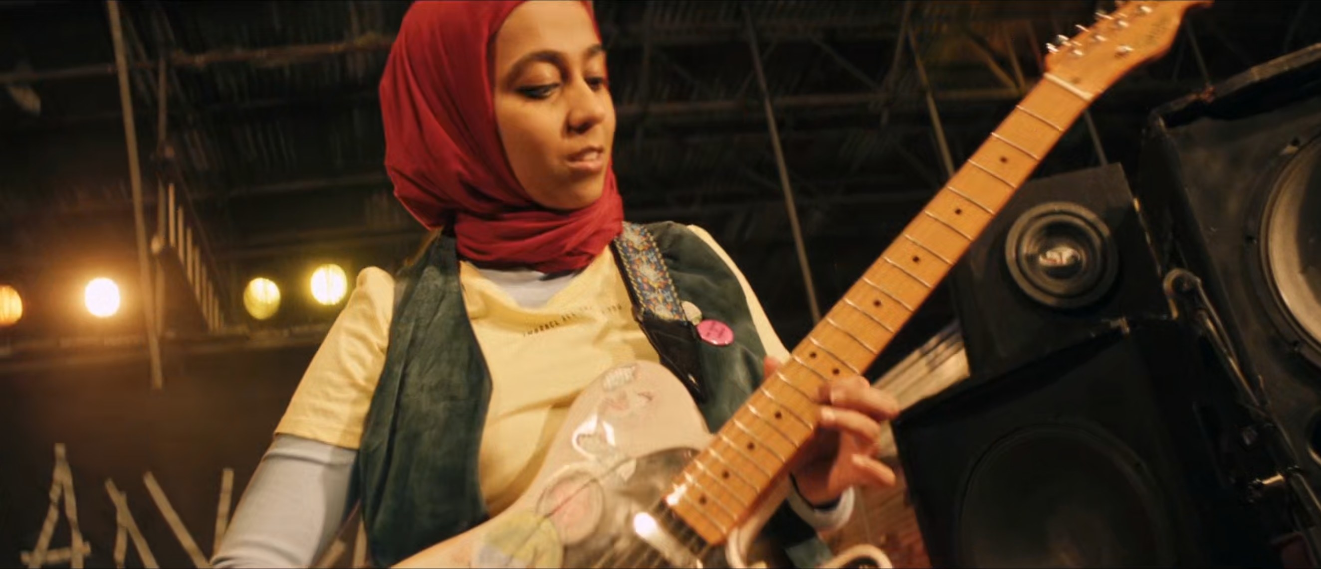 In a screenshot from the season 1 finale, Amina, a Muslim woman wearing a red hijab, looks content on stage playing electric guitar with her band, who are not in frame.