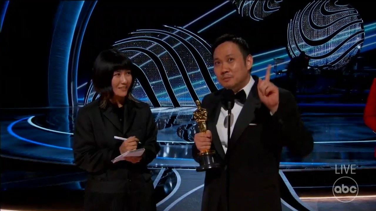 Ryusuke Hamaguchi onstage holding his Oscar, with a woman standing next to him