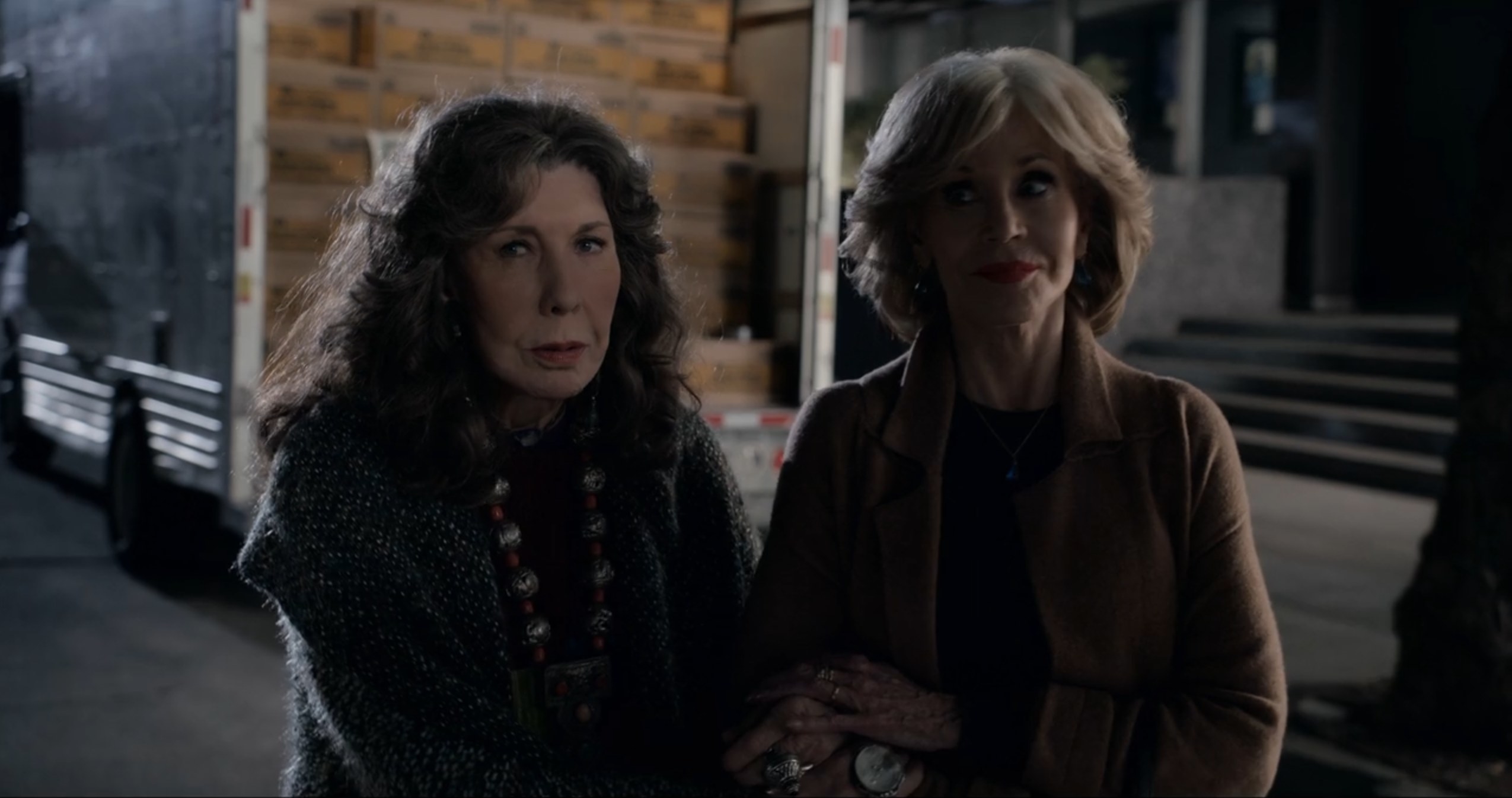 A screenshot of Grace and Frankie walking arm-in-arm. It is hark outside and Grace is smiling, Frankie is talking. They are both looking to the right.