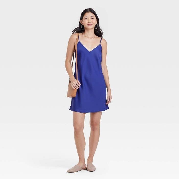Model wearing the blue thigh-length spaghetti strap slip dress with shoulder purse and flats
