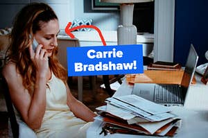 Carrie Bradshaw, while talking on a landline phone, sits in front of a messy desk filled with papers and laptops