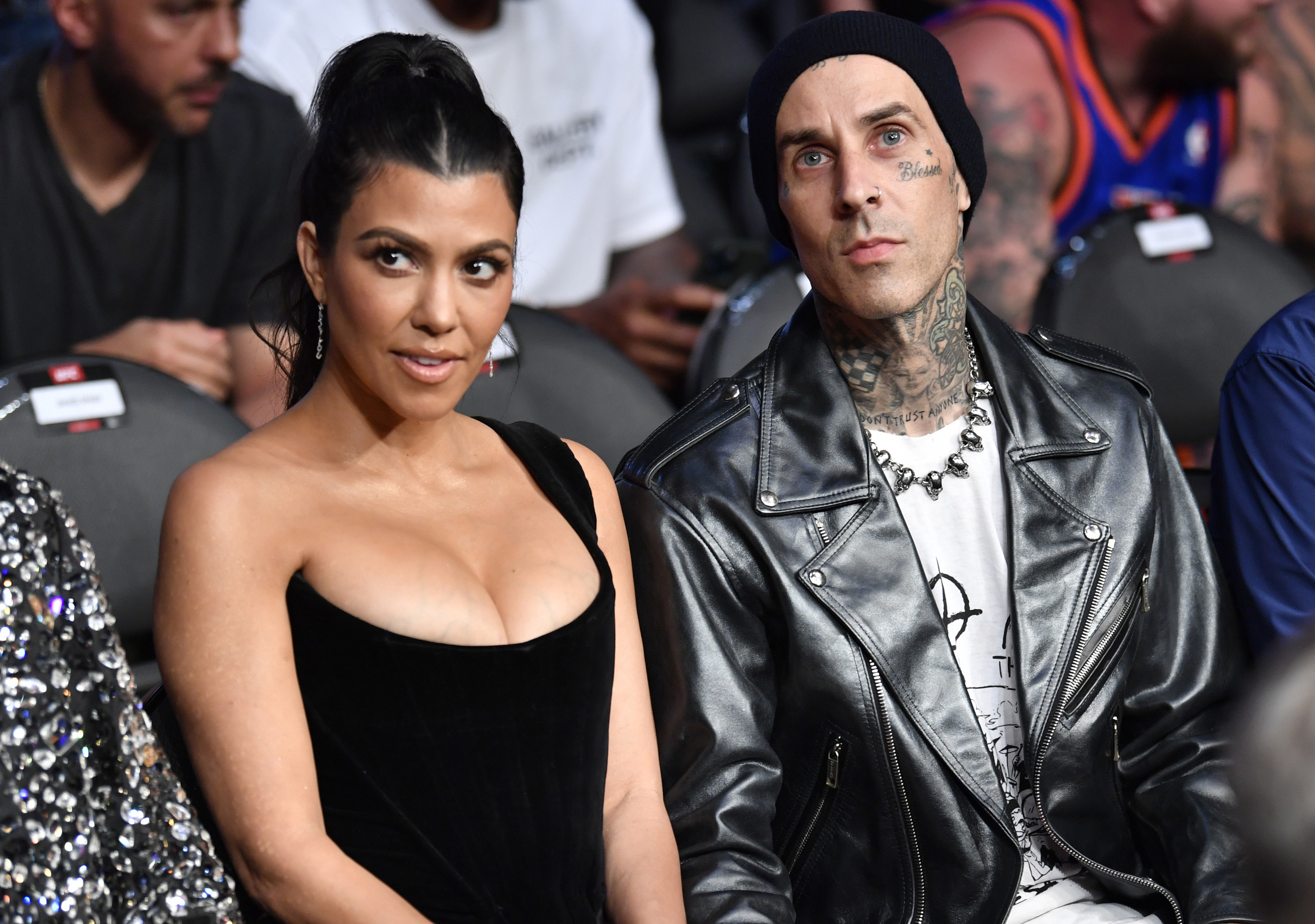 Kourtney in a one-strapped dress sits next to fiance Travis Barker wearing a leather jacket and beanie