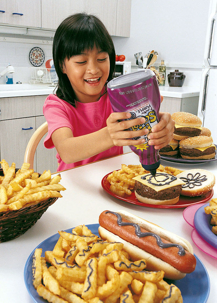A little girl happily squeezes purple EZ Squirt ketchup onto burgers, fries, and hot dogs