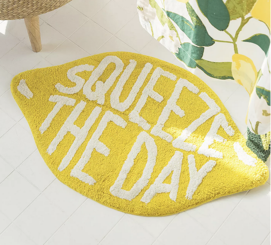 The lemon-shaped bath mat embroidered with the words, &quot;Squeeze the Day&quot;, on a floor next to a shower curtain with lemon tree designs