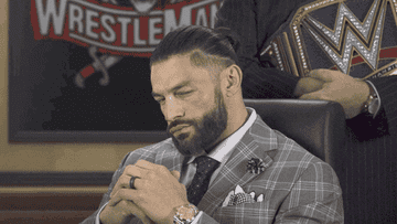 Roman Reigns in a suit, sits all smug