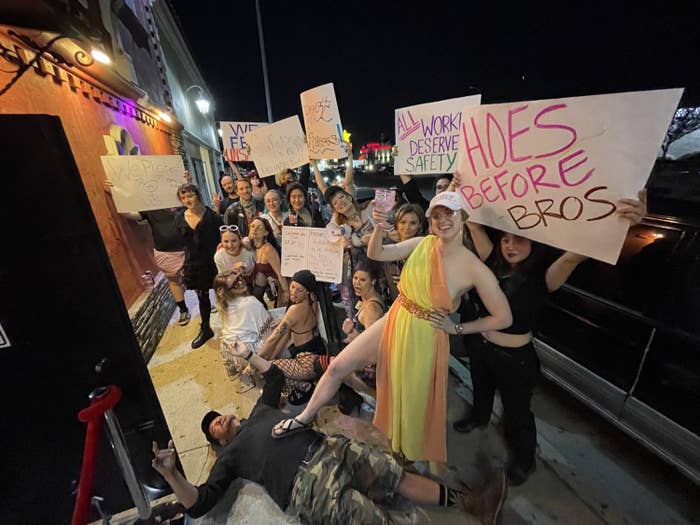 A group of people with hold signs that read "all workers deserve safety" and "hoes before bros" in protest at a Hollywood strip club