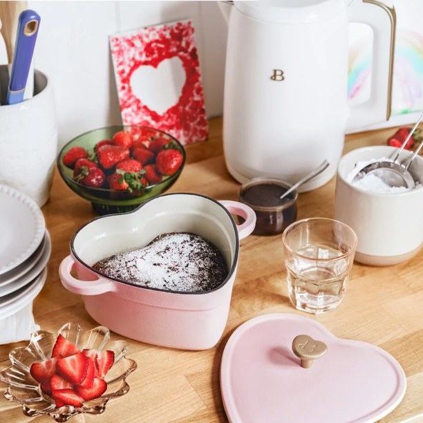 Pink heart shaped dutch oven with chocolate cake inside, next to heart shaped lid, on table with strawberries and glassware