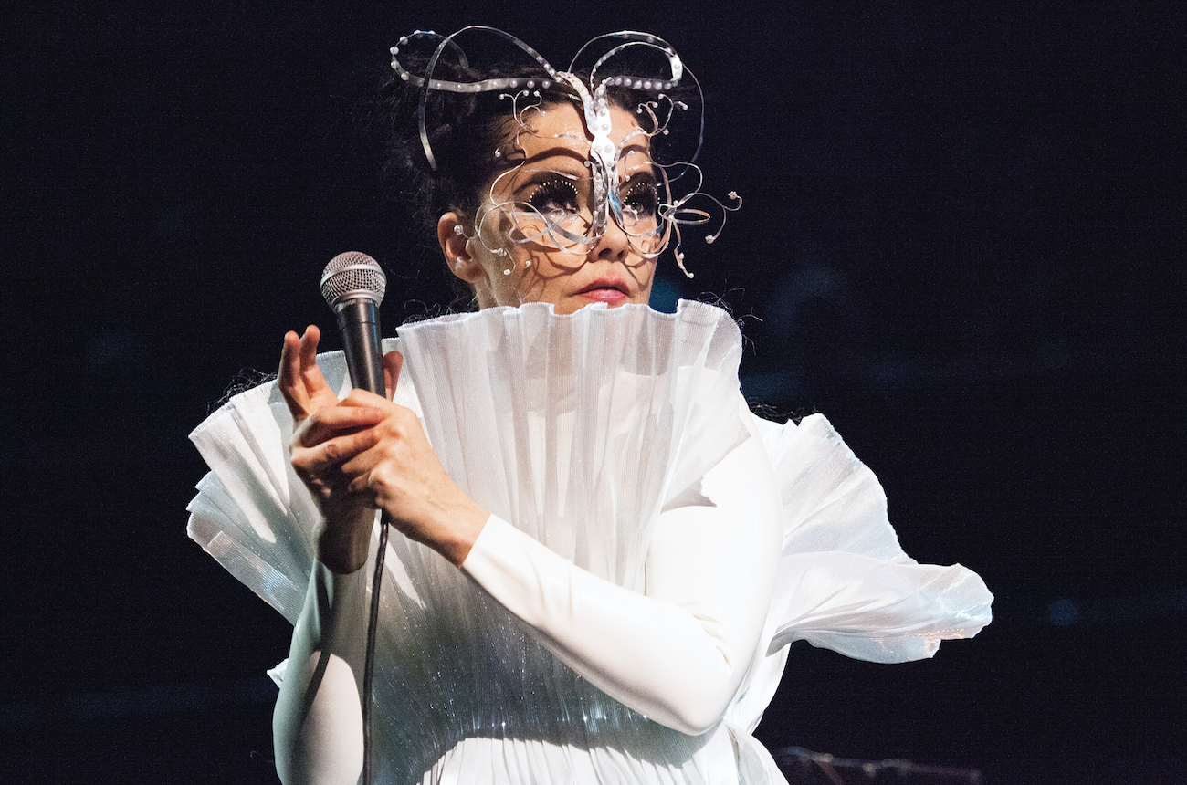 Bjork holds the mic away from her face wearing head/face jewelry piece and a pleated dress