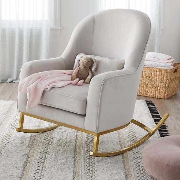 Beige rocking chair with gold rocker on the bottom, pink blanket and teddy bear on it