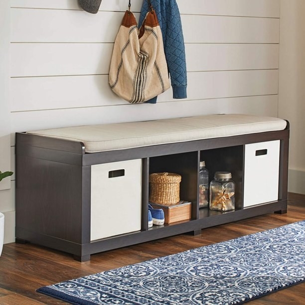 Espresso wood colored storage bench with beige bench cushion, two white cubes on left and right, open cubbies in the middle with objects inside