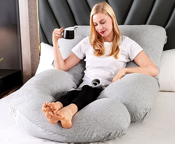 A person on their bed sitting on the pillow while they drink coffee and read a magazine