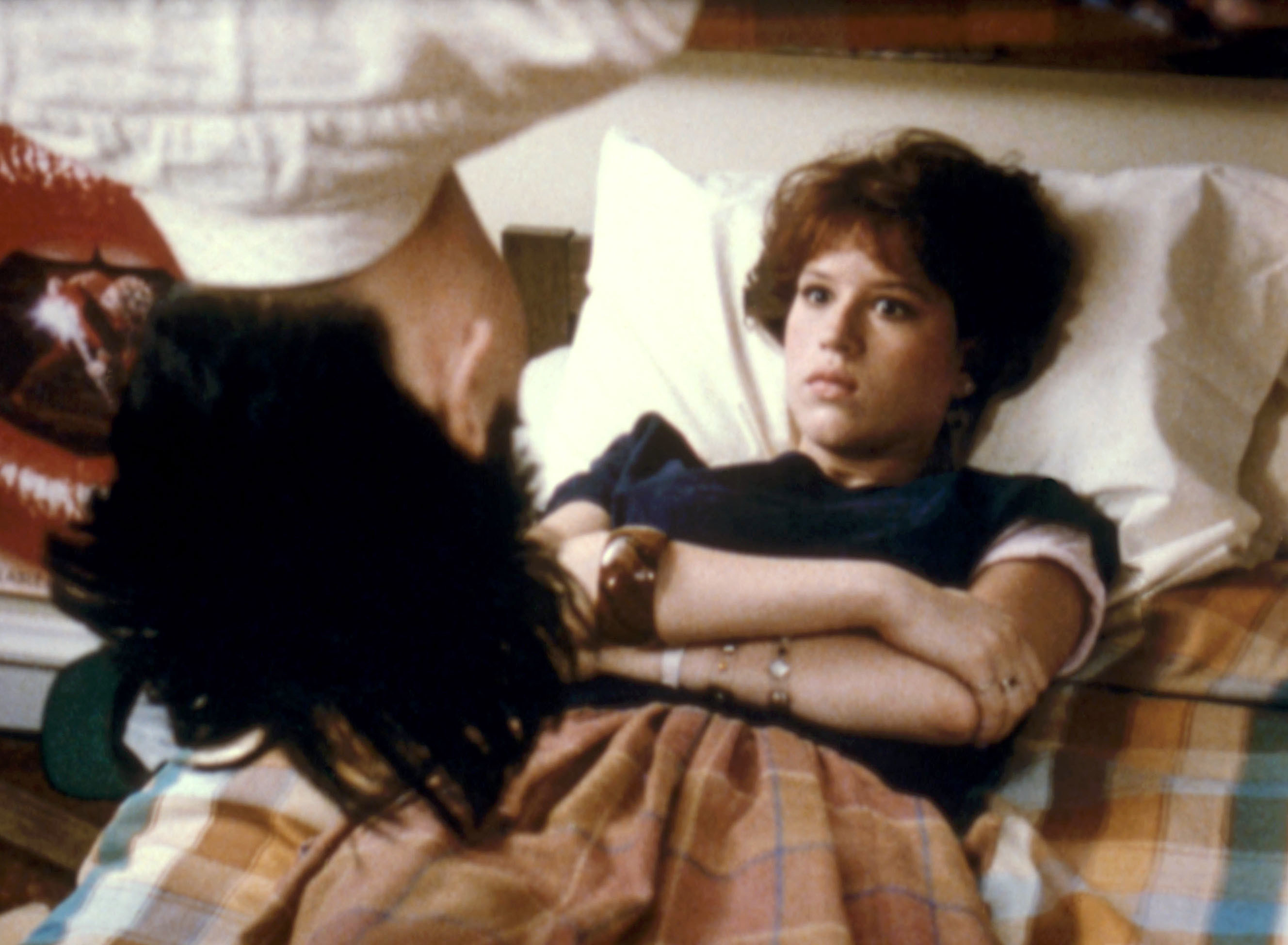 Molly Ringwald lays in bed as a person hands upside down in front of her