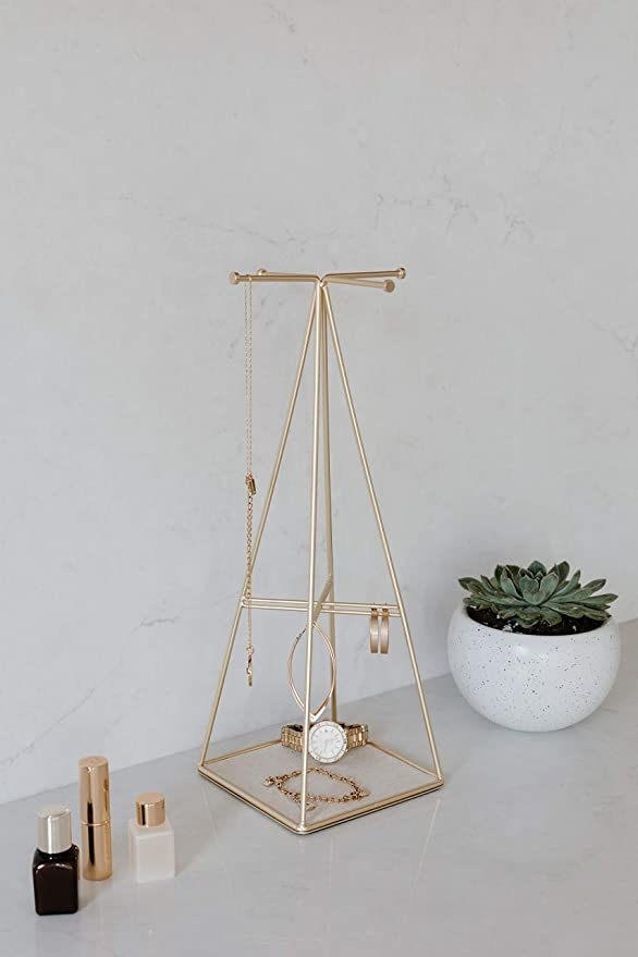 The jewellery stand on a counter next to a plant and cosmetic bottles