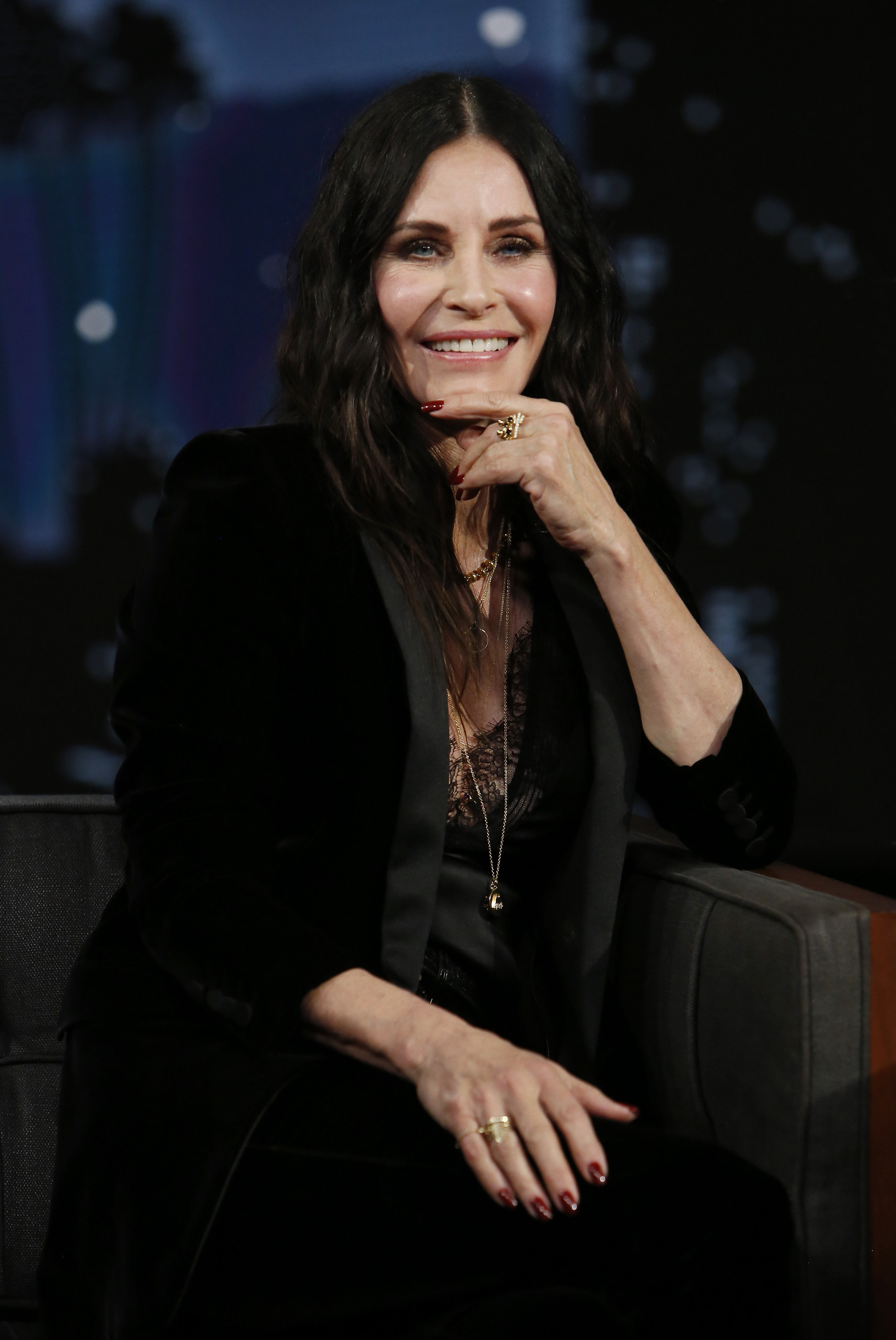 Courtney Cox sits on a couch for a late night talk show, wearing a black suit and her hand on her chin