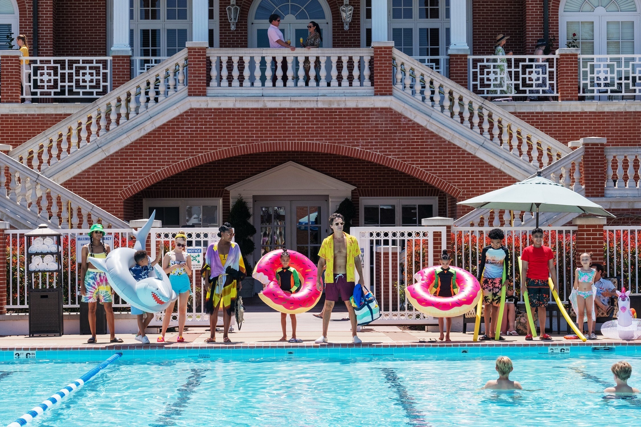 A group of adults (Gabrielle Union and Zach Braff) and children stand by the side of a pool holding different floaties
