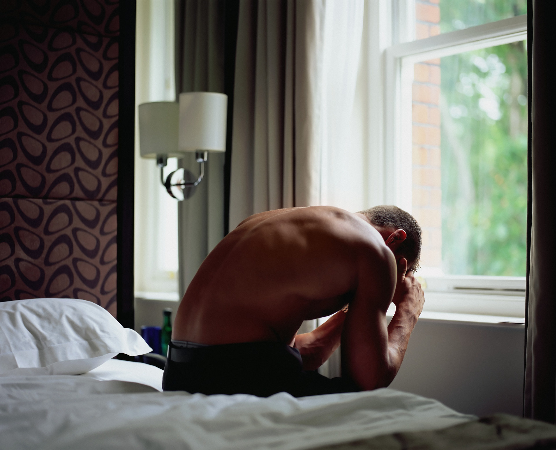 Shirtless man sitting at the edge of bed leaning over in despair