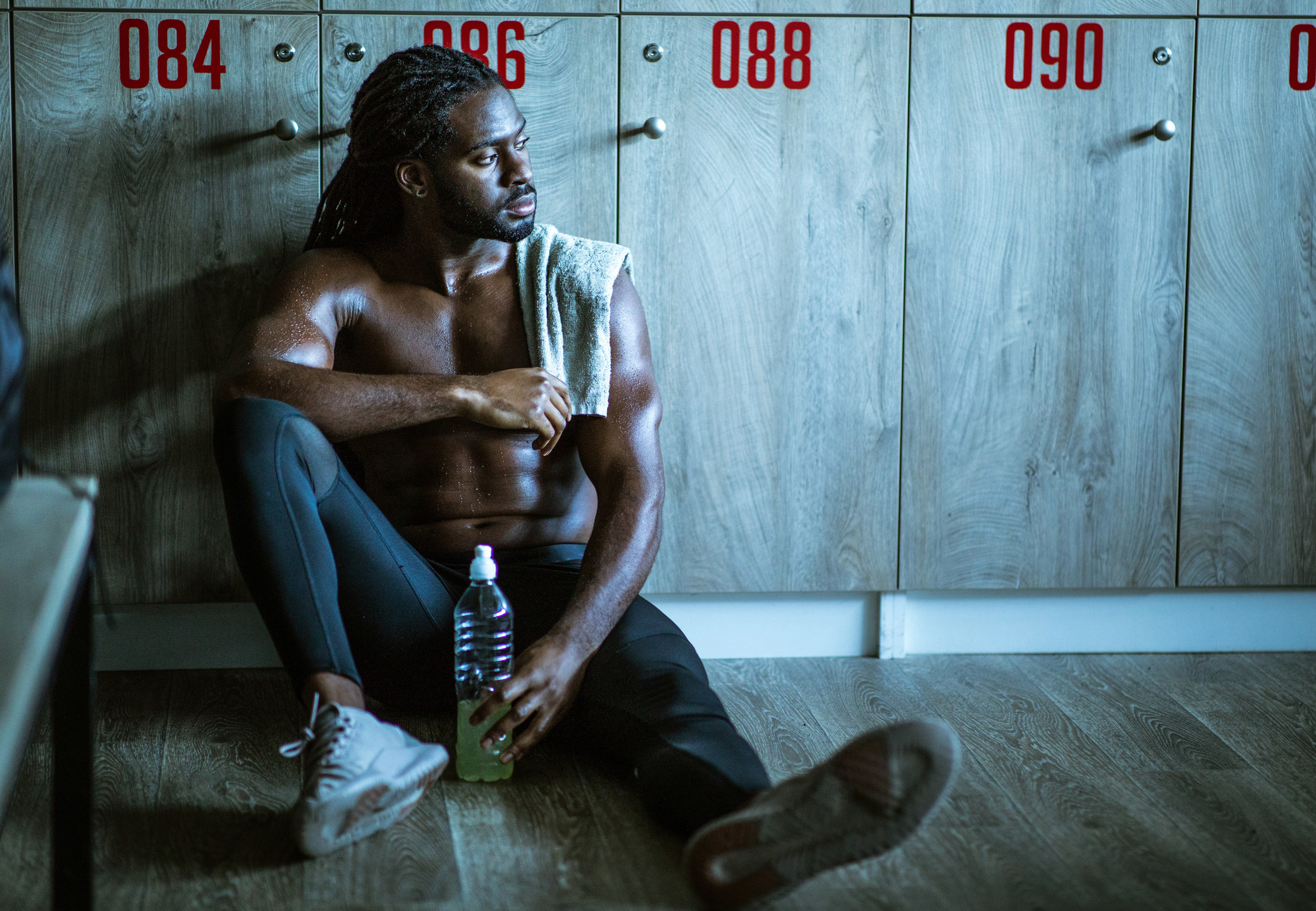 Shirtless athlete sitting on floor of locker room looking pensive