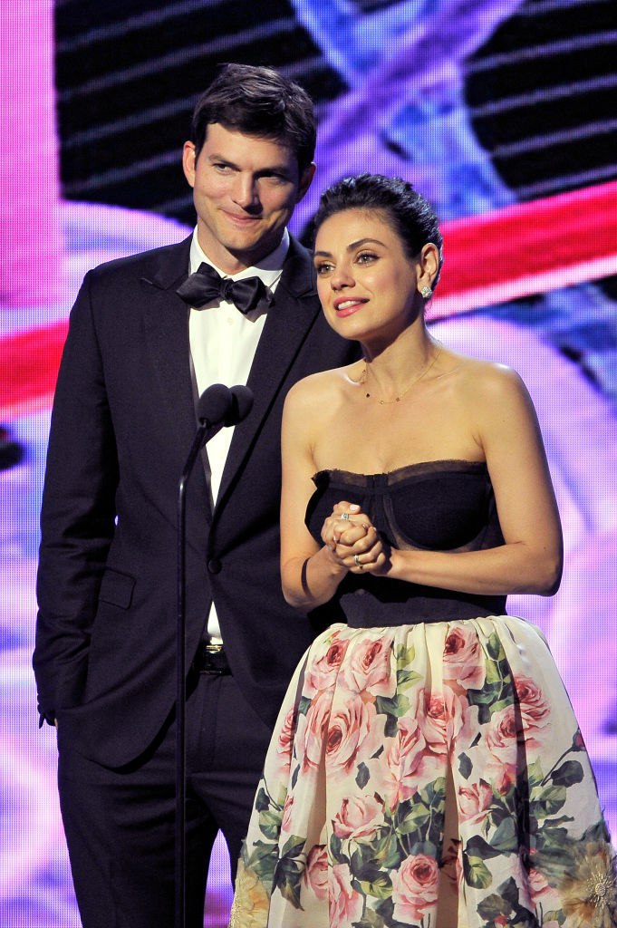 Mila and Ashton stand onstage for an award show, Mila wears a strapless black and floral dress while Ashton wears a suit