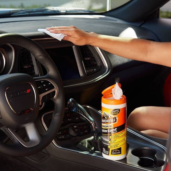 a model using a wipe on a black dashboard, with the orange container of wipes on the center console