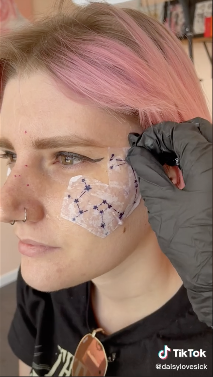 Screengrab from a TikTok by user daisylovesick of a hand placing a tattoo stencil of a constellation on a client&#x27;s face