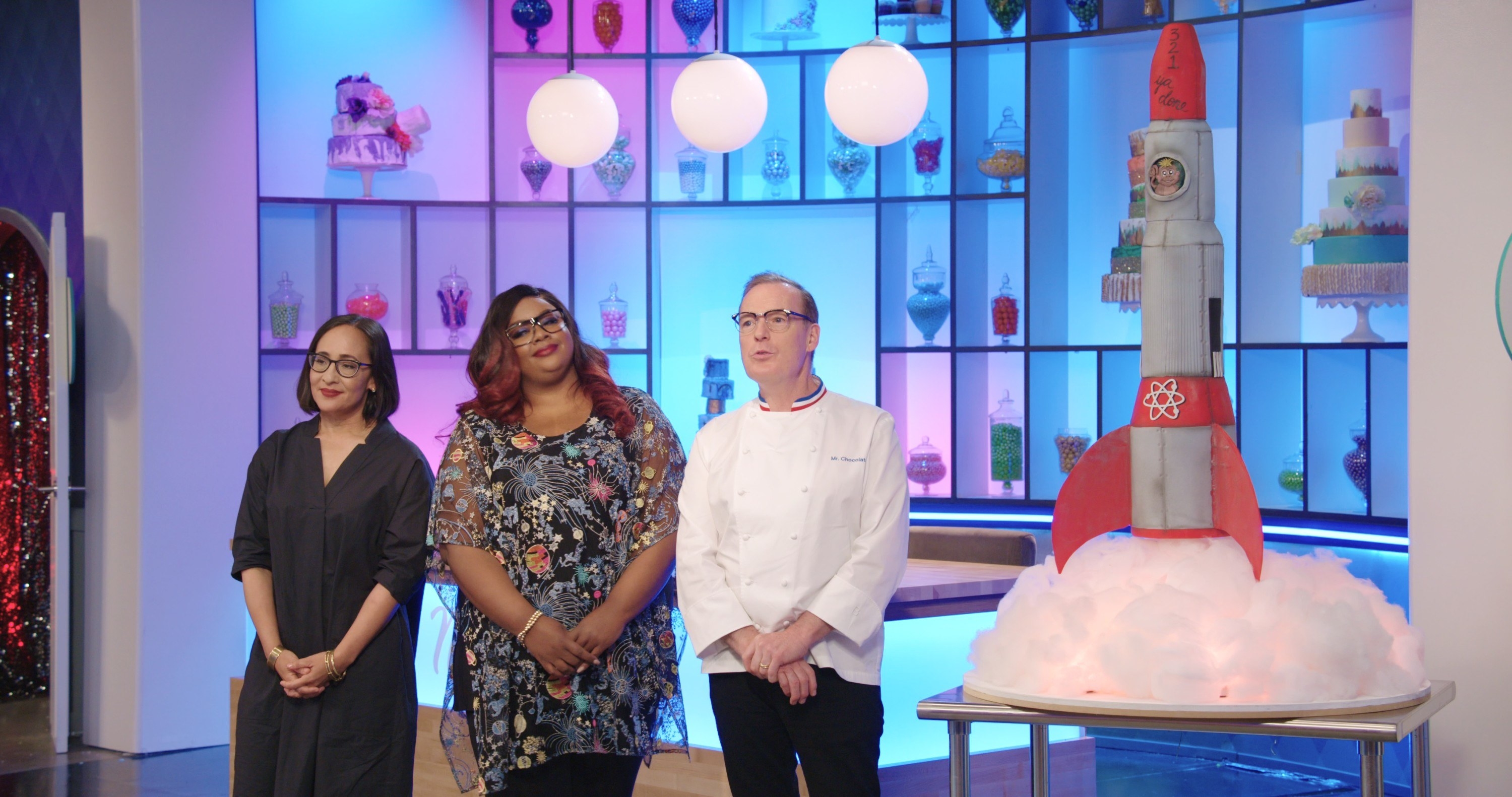 the judges of Nailed It next to a rocketship cake