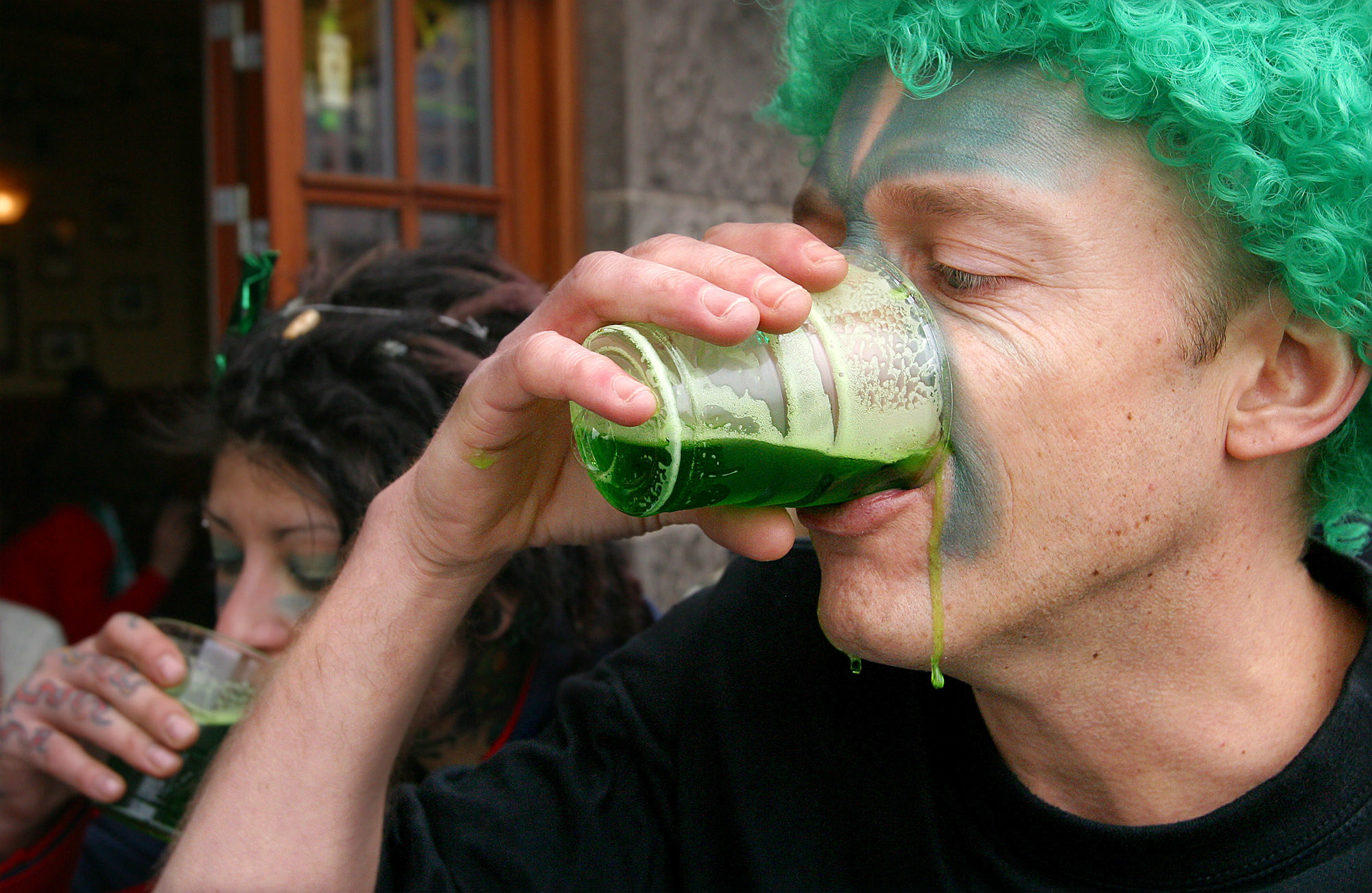 a man in a green wig chugs green beer with some of it dripping, a woman drinks green beer behind him 