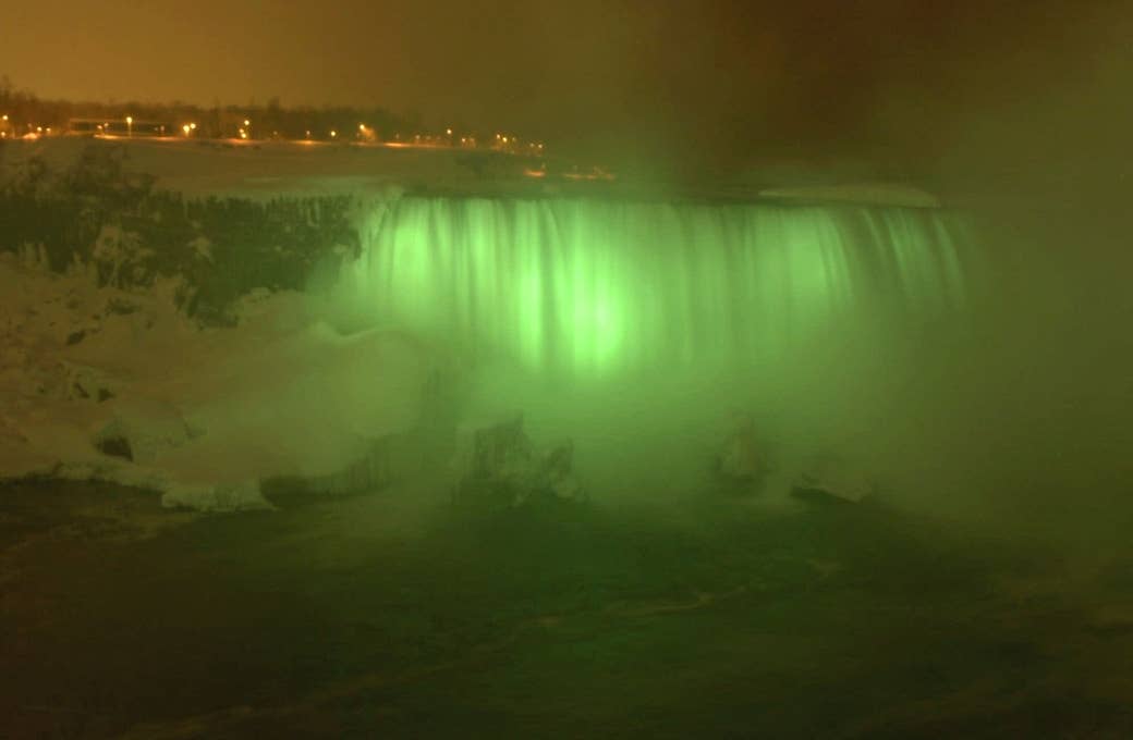 Niagara Falls at night with mist and green lights that make it look green, with a town in the background 